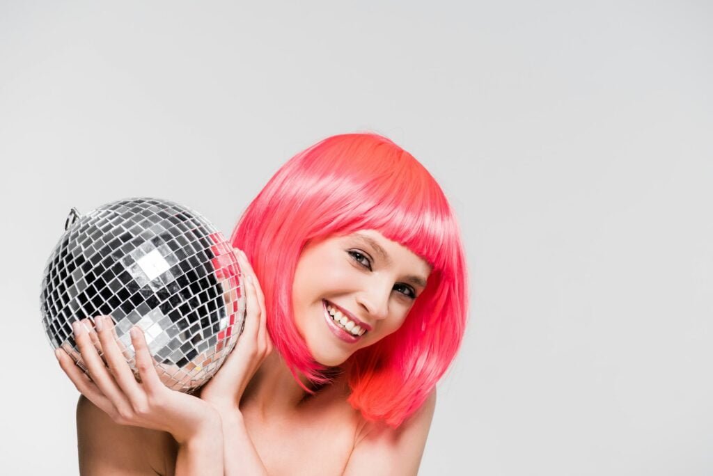 smiling girl in pink wig holding disco ball, isolated on grey